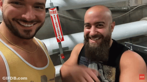 Two Men Smiling In Front Of A Fire Extinguisher GIF