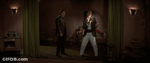 Two Men Standing In A Living Room With A Green Curtain GIF