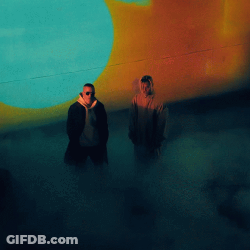 Two Men Standing In Front Of A Wall With A Blue And Orange Circle On It GIF