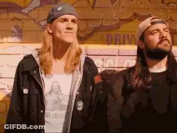 Two Men Standing Next To Each Other In Front Of A Wall With Graffiti On It That Says Drive GIF