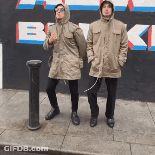 Two Men Standing On A Sidewalk In Front Of A Wall That Has The Letter B Painted On It GIF