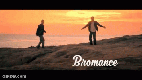 Two Men Standing On Top Of A Sandy Hill With The Word Bromance Written Below Them GIF