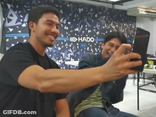 Two Men Take A Selfie In Front Of A Wall That Says Hado Blue GIF