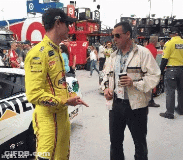 Two Men Talking In Front Of A Race Car That Says Pennzoil GIF