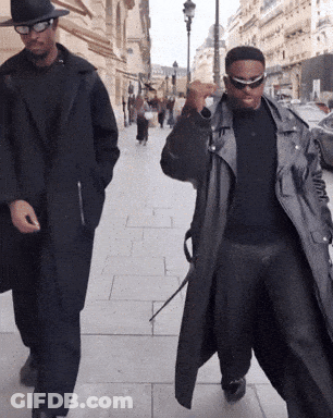 Two Men Walking Down A Sidewalk One Wearing A Hat And Sunglasses GIF