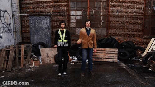 Two Men Walking Past Brick Building With Graffiti GIF