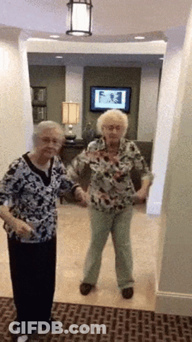 Two Older Women Are Dancing In A Hallway In Front Of A Tv GIF