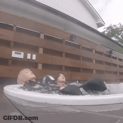 Two People Are Laying In A Hot Tub Outside A House With A Clock On The Wall GIF