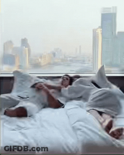 Two People Are Laying On A Bed In Front Of A Window With A City In The Background GIF