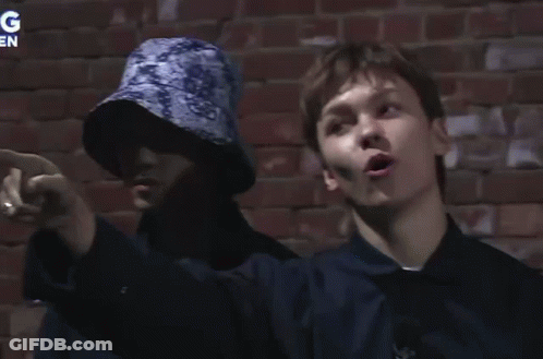 Two People Are Standing In Front Of A Brick Wall With The Letter G On The Bottom Right GIF