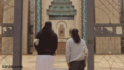 Two People Are Standing In Front Of A Building With Arabic Writing On The Wall GIF