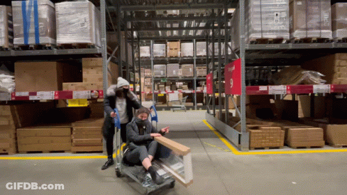 Two People Pushing A Cart In A Warehouse With A Red Sign That Says Ikea GIF