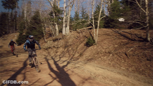 Two People Riding Bikes Down A Dirt Road GIF