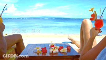 Two People Sit At A Table On A Beach Holding Drinks GIF