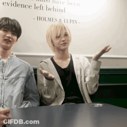 Two People Sitting At A Table With A Sign That Says Holmes & Lupin GIF