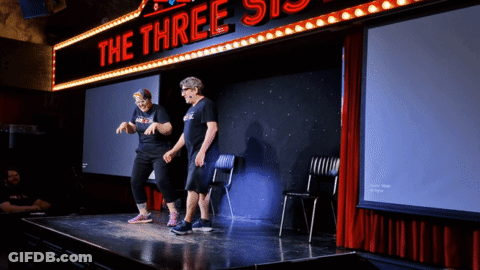 Two People Stand On A Stage Under A Neon Sign That Says The Three Sisters GIF