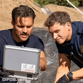 Two Police Officers Are Looking At A Case With A Warning Sticker On It GIF