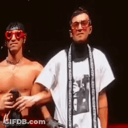 Two Shirtless Men Are Standing Next To Each Other On A Stage . One Of The Men Is Wearing Sunglasses And A Scarf . GIF