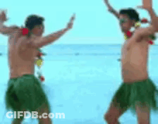 Two Shirtless Men In Hula Skirts Dancing On Beach GIF