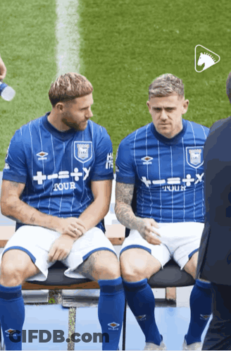 Two Soccer Players Are Sitting On A Bench And One Has A Shirt That Says Tour GIF