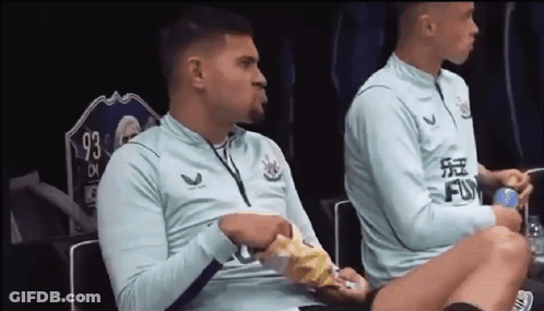 Two Soccer Players Are Sitting On A Bench In A Locker Room Eating Chips . GIF