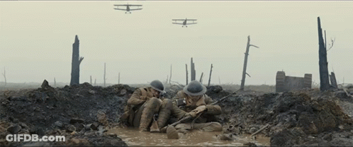 Two Soldiers Are Laying In A Muddy Trench With Planes Flying In The Background GIF