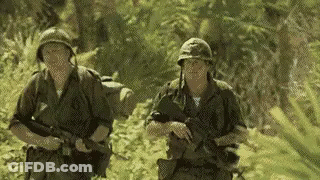 Two Soldiers Are Walking Through A Lush Green Forest . GIF