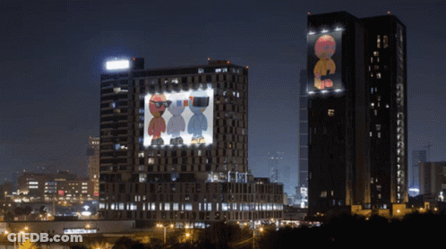 Two Tall Buildings Are Lit Up At Night And One Has A Picture Of Two Cartoon Characters Projected On It GIF