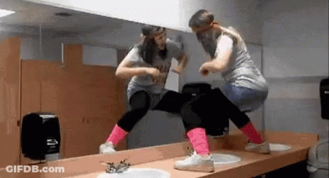 Two Women Are Dancing In Front Of A Mirror In A Public Restroom . GIF