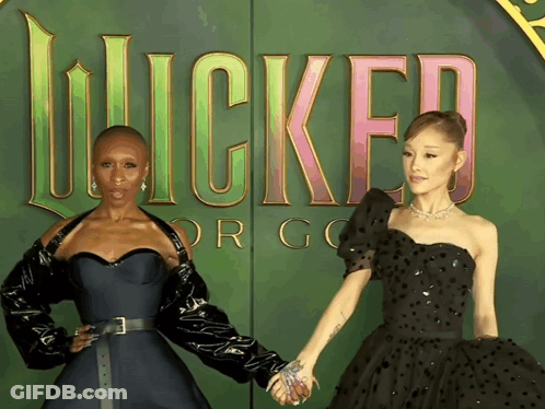 Two Women Are Holding Hands In Front Of A Sign That Says Wicked GIF