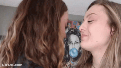 Two Women Are Kissing In Front Of A Painting Of A Woman Blowing A Blue Bubble Gum . GIF