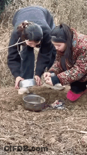 Two Women Are Kneeling Down In The Dirt Looking At A Bowl Of Food . GIF