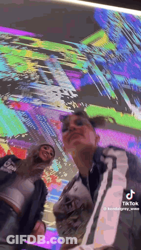 Two Women Are Posing For A Picture In Front Of A Large Screen . GIF