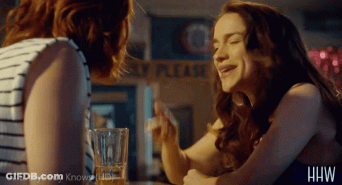 Two Women Are Sitting At A Table In Front Of A Sign That Says " Please " GIF