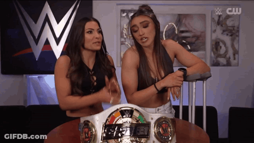 Two Women Are Sitting At A Table In Front Of A Wwe Sign GIF