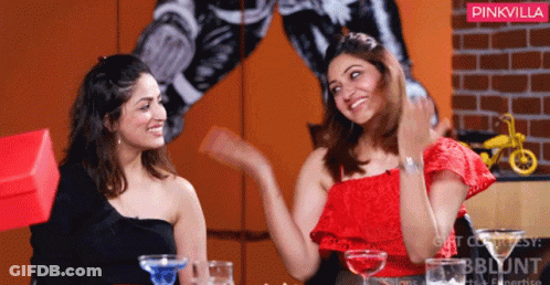 Two Women Are Sitting At A Table With Drinks In Front Of A Wall With A Pinkvilla Logo GIF