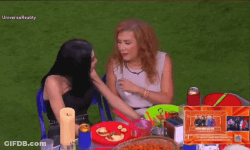 Two Women Are Sitting At A Table With Plates Of Food And The Word Universoreality On The Bottom Right GIF