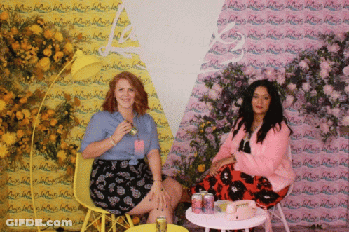 Two Women Are Sitting In Front Of A Wall That Has La Croix Written On It GIF