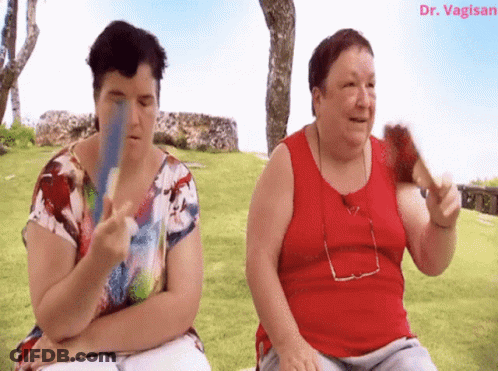 Two Women Are Sitting In The Grass And One Is Holding A Fan And The Other Is Holding A Card GIF