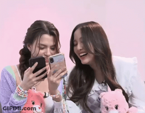 Two Women Are Sitting Next To Each Other And Looking At Their Phones . GIF