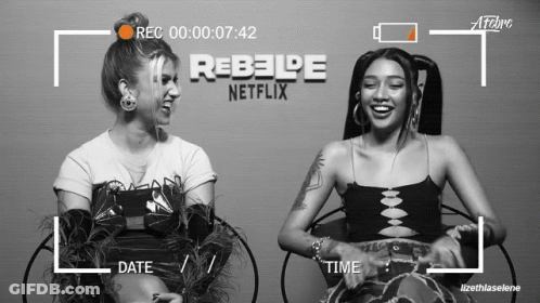 Two Women Are Sitting Next To Each Other In Front Of A Rebelde Netflix Logo GIF
