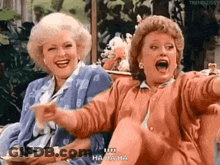 Two Women Are Sitting Next To Each Other Laughing And Pointing At Something . GIF