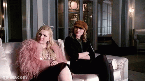 Two Women Are Sitting On A Couch And One Of Them Is Wearing A Pink Fur Coat . GIF