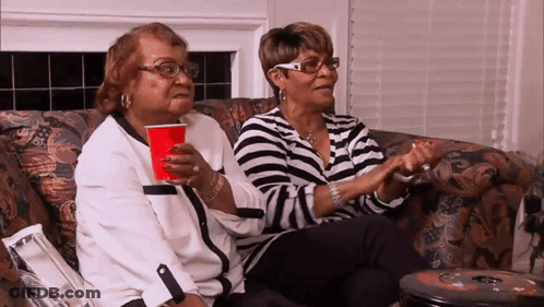 Two Women Are Sitting On A Couch Holding Hands And One Is Holding A Red Cup GIF