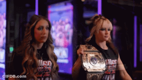 Two Women Are Standing Next To Each Other And One Has A Wrestling Championship Belt GIF