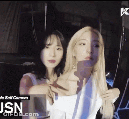 Two Women Are Standing Next To Each Other In A Dark Room . One Of The Women Is Taking A Selfie With Her Phone . GIF