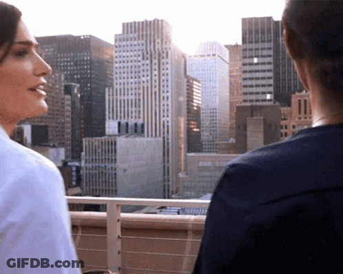 Two Women Are Standing On A Balcony Overlooking A City . GIF