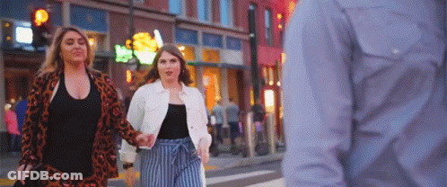 Two Women Are Walking Down A City Street Holding Hands While A Man Looks On . GIF