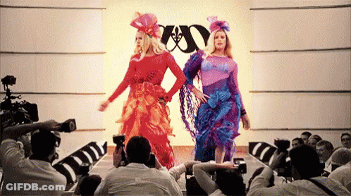 Two Women Are Walking Down A Runway At A Fashion Show While A Crowd Watches . GIF