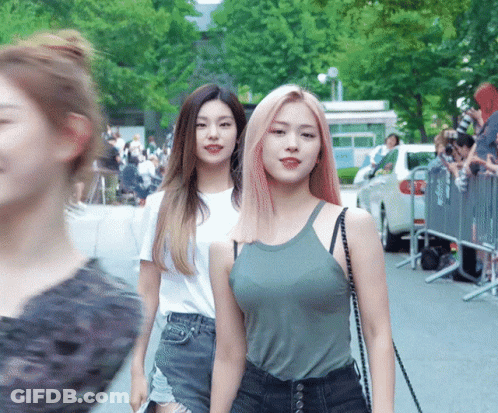 Two Women Are Walking Down A Street With A Crowd Behind Them GIF